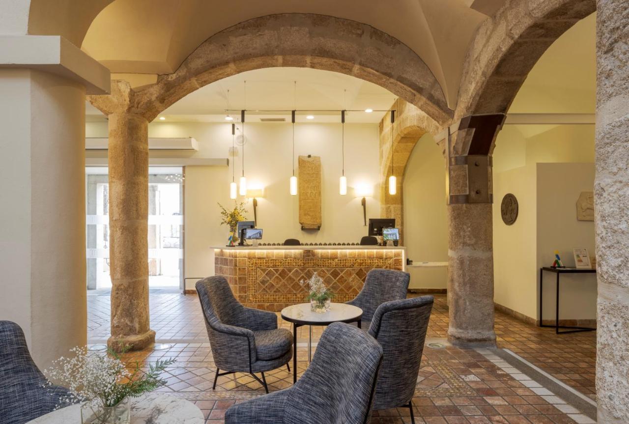 Hotel Ilunion Merida Palace Экстерьер фото The photo shows the interior of a hotel or a similar establishment. It features a spacious lobby area with an arched ceiling and stone columns. The reception desk is located at the back, designed with a textured surface and illuminated by pendant lig